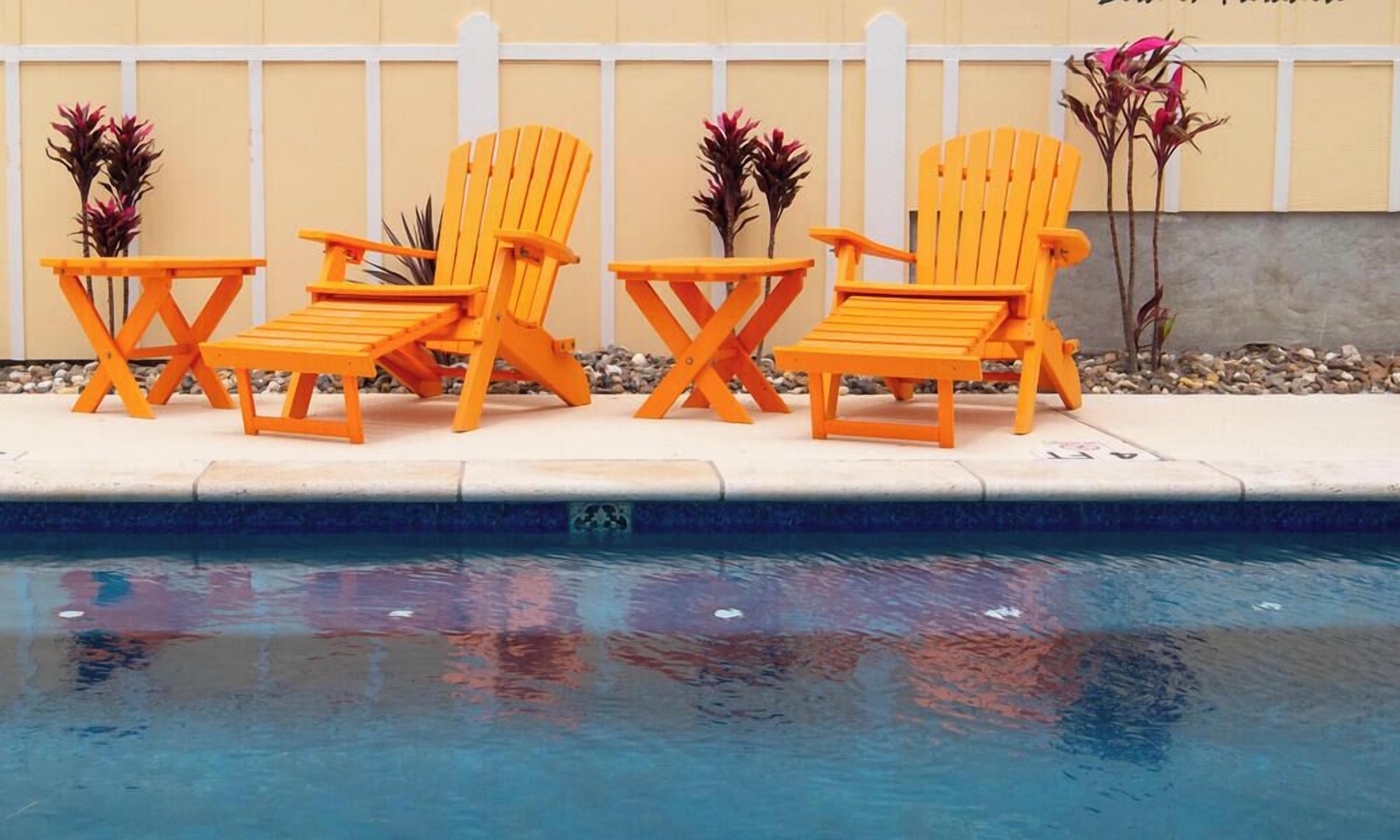 Un área junto a la piscina con tres sillas Adirondack naranja y un letrero que dice 'Pelicans Roast #103 Bird in Paradise'.