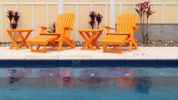 Un área junto a la piscina con tres sillas Adirondack naranja y un letrero que dice 'Pelicans Roast #103 Bird in Paradise'.