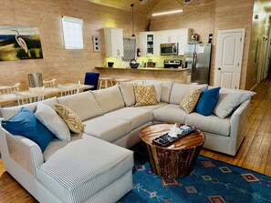 A modern living room with a sectional sofa, a coffee table, and a kitchen area with white cabinets and appliances. - Hot tub, Pool, blackouts, 75" TV, Dogs OK (Port Aransas)