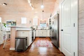 A compact kitchen with wooden flooring, white cabinets, a stainless steel refrigerator, and a small trash bin.
