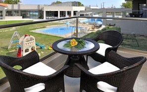 A balcony with wicker chairs and a table, overlooking a swimming pool and a children's play area.