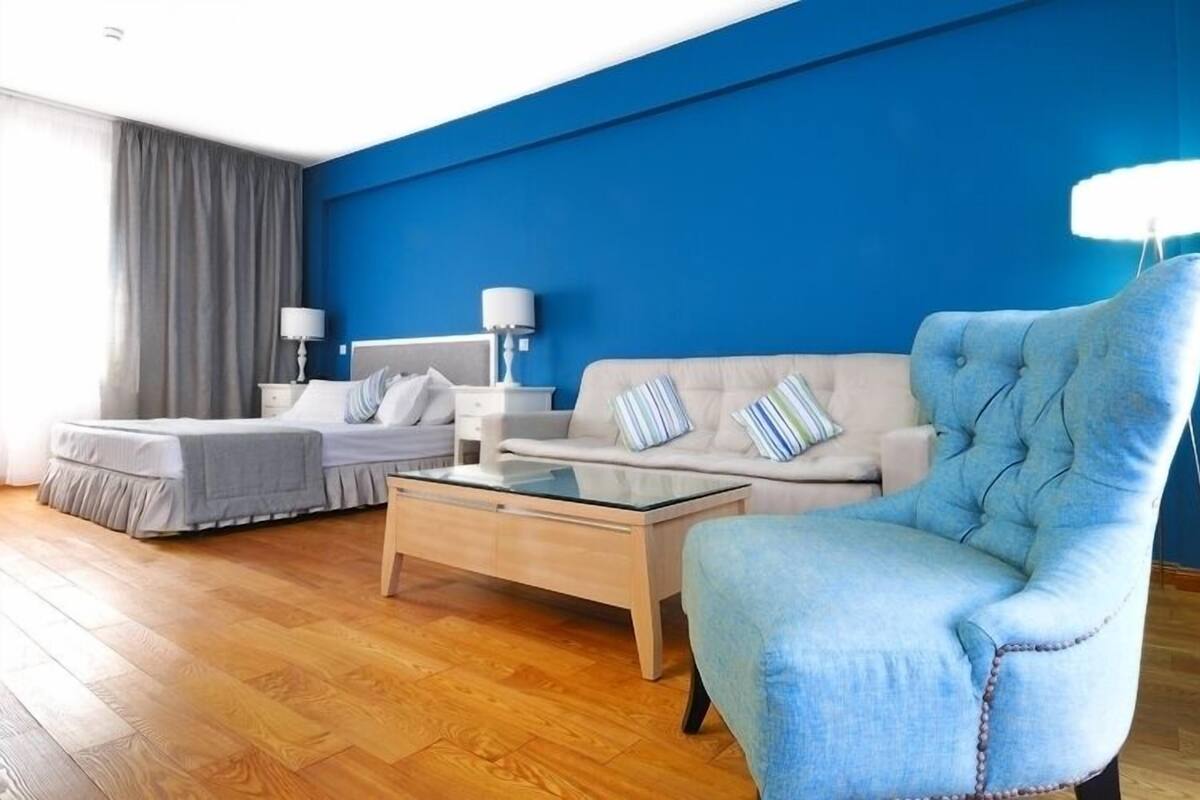 Ein modernes Hotelzimmer mit blauer Akzentwand, einer Couch, einem Sessel, einem Bett und einem Couchtisch.