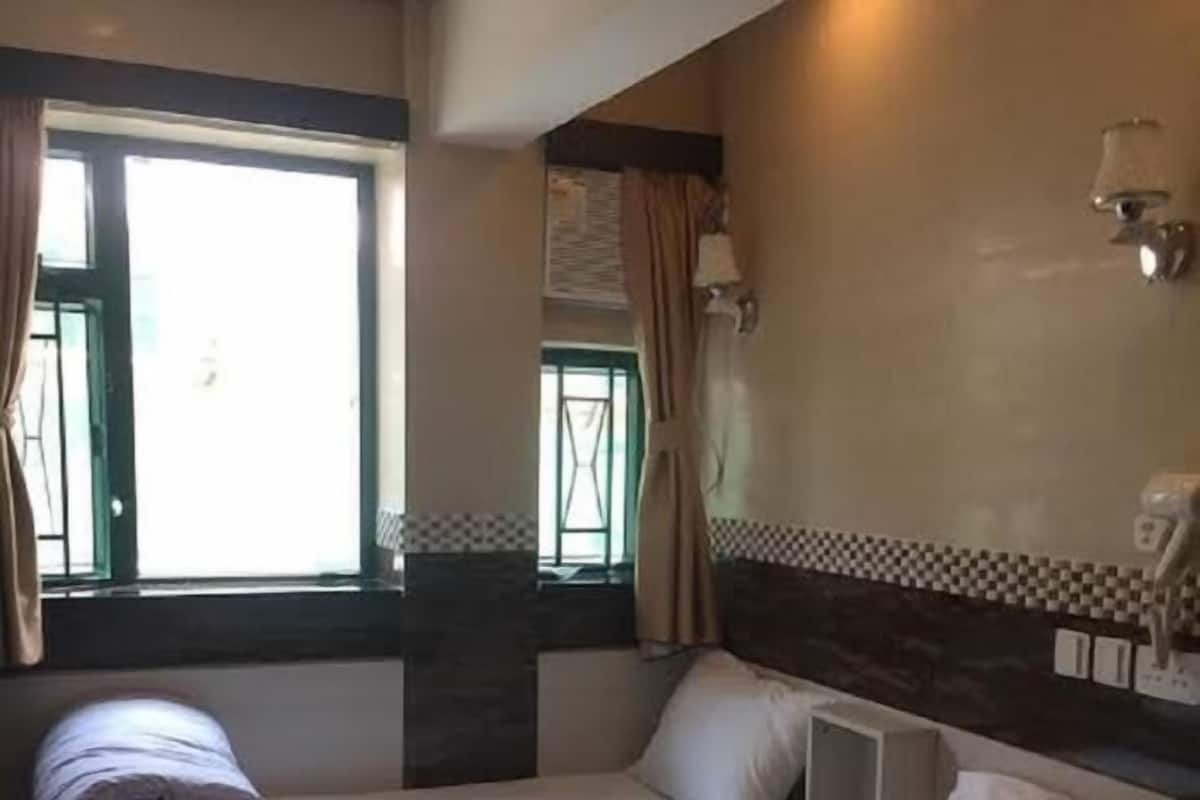 A hotel room with two single beds, a small table, a window with curtains, and a wall-mounted light fixture.