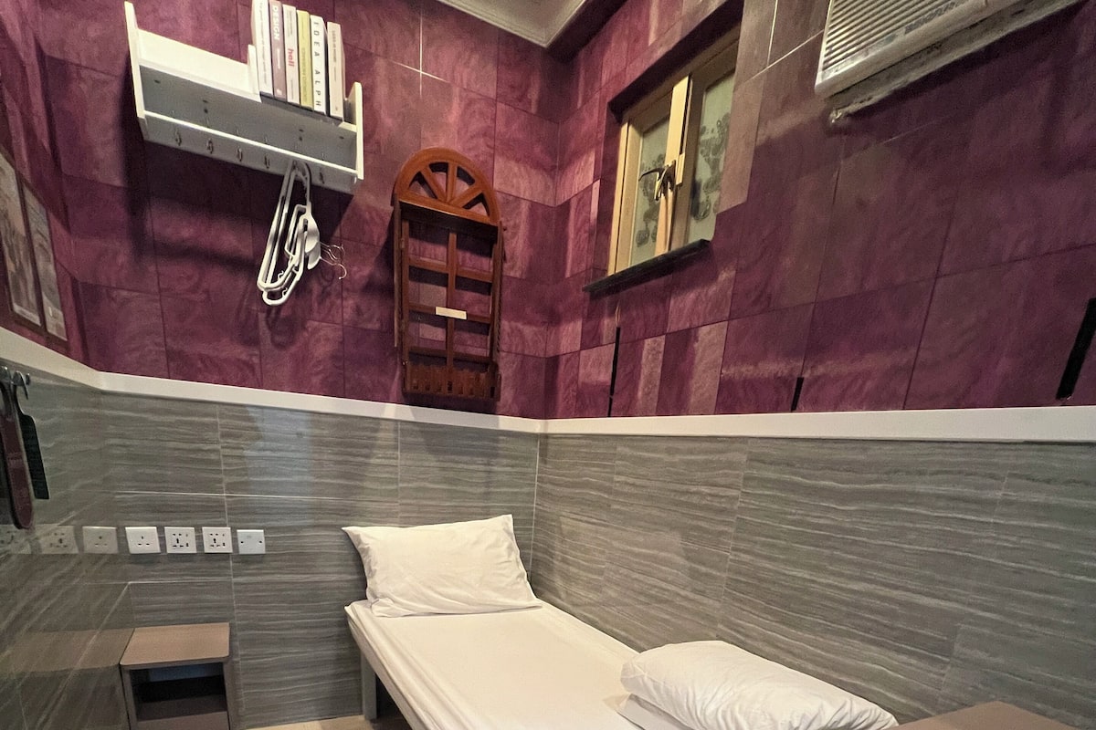 A compact hotel room with a single bed, a small bedside table, a built-in shelf with books, a wooden chair, and a small trash bin.