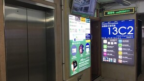Un ascensor amb un panell digital que mostra el número 13C2. Hi ha senyals per a la recepció d'un hostal i una zona de control de seguretat.