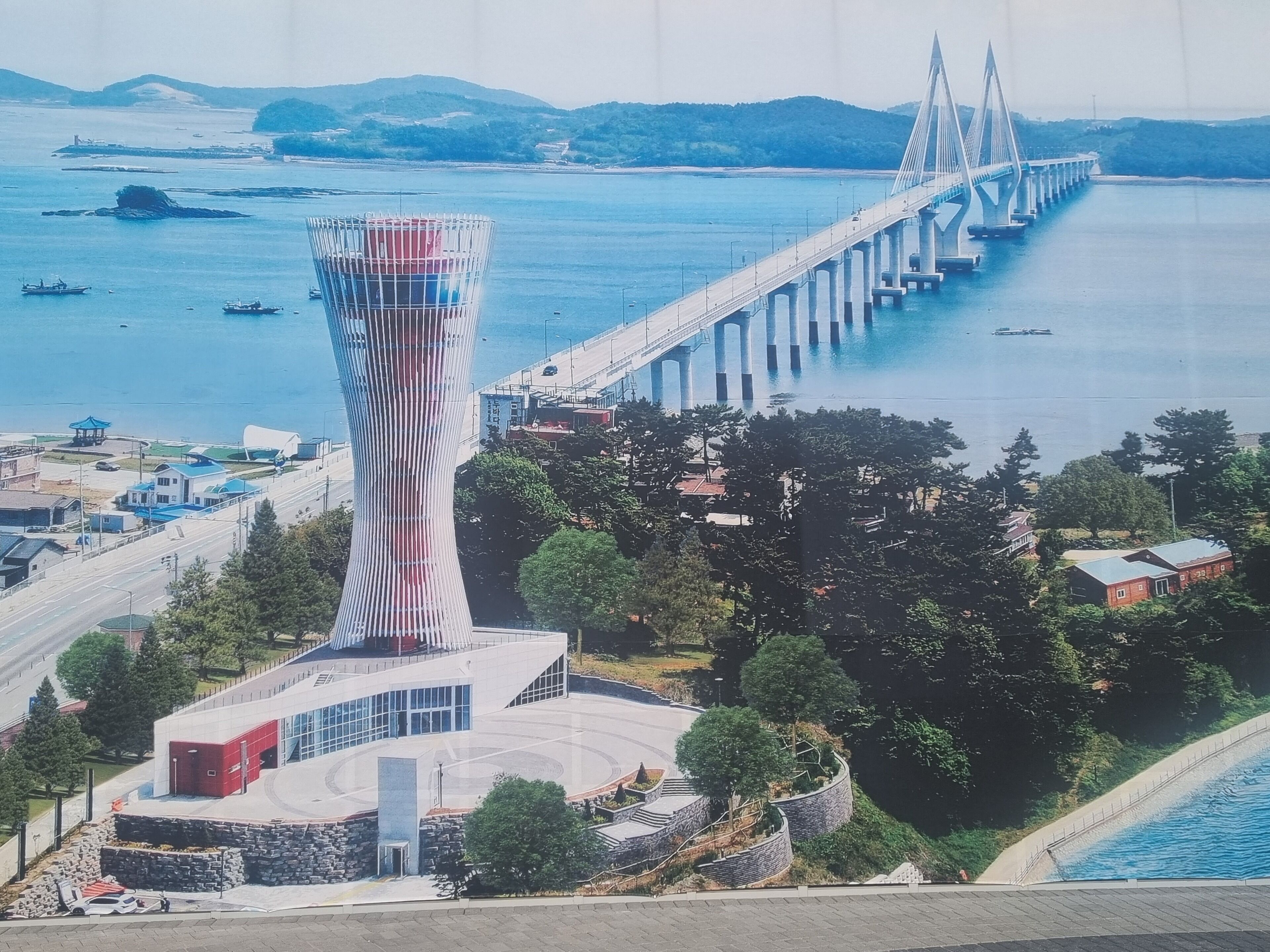 A modern tower with a unique twisted design, a bridge in the background, and a coastal setting with boats and greenery.