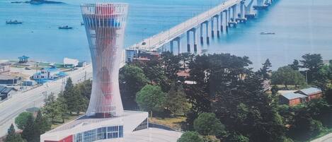 A modern tower with a unique twisted design, a bridge in the background, and a coastal setting with boats and greenery.