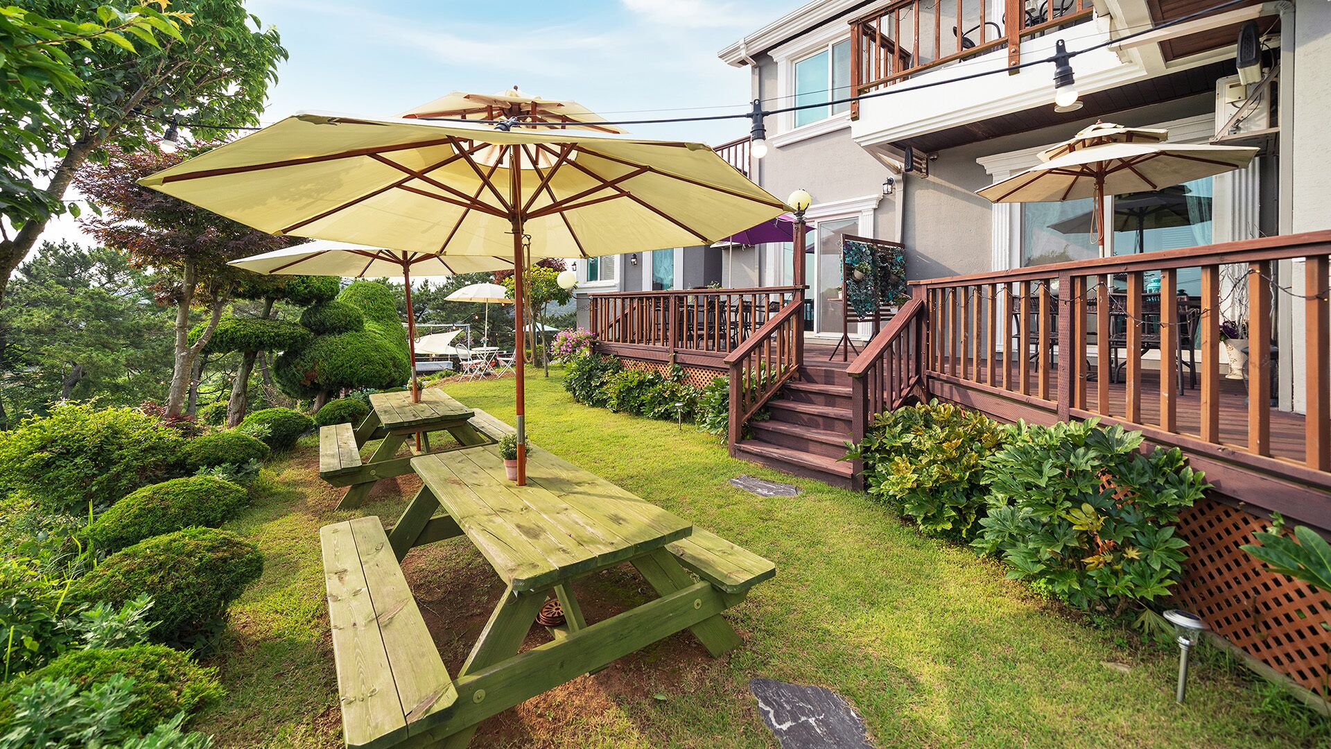 Eine Terrasse mit Picknicktischen und Sonnenschirmen, umgeben von gepflegten Büschen und einer kleinen Rasenfläche.