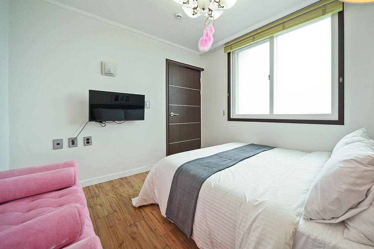 Ein Schlafzimmer mit einem Bett, einem an der Wand befestigten Fernseher, einem Fenster mit Jalousien, einem rosa Sessel und Holzboden.