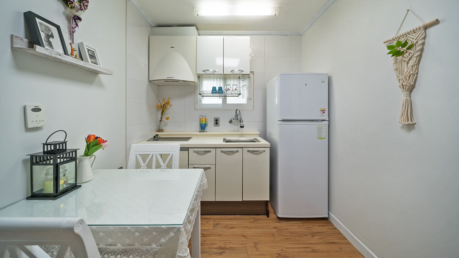 Una cocina compacta con armarios blancos, un refrigerador blanco, un lavamanos pequeño y una mesa de comedor con mantel blanco.
