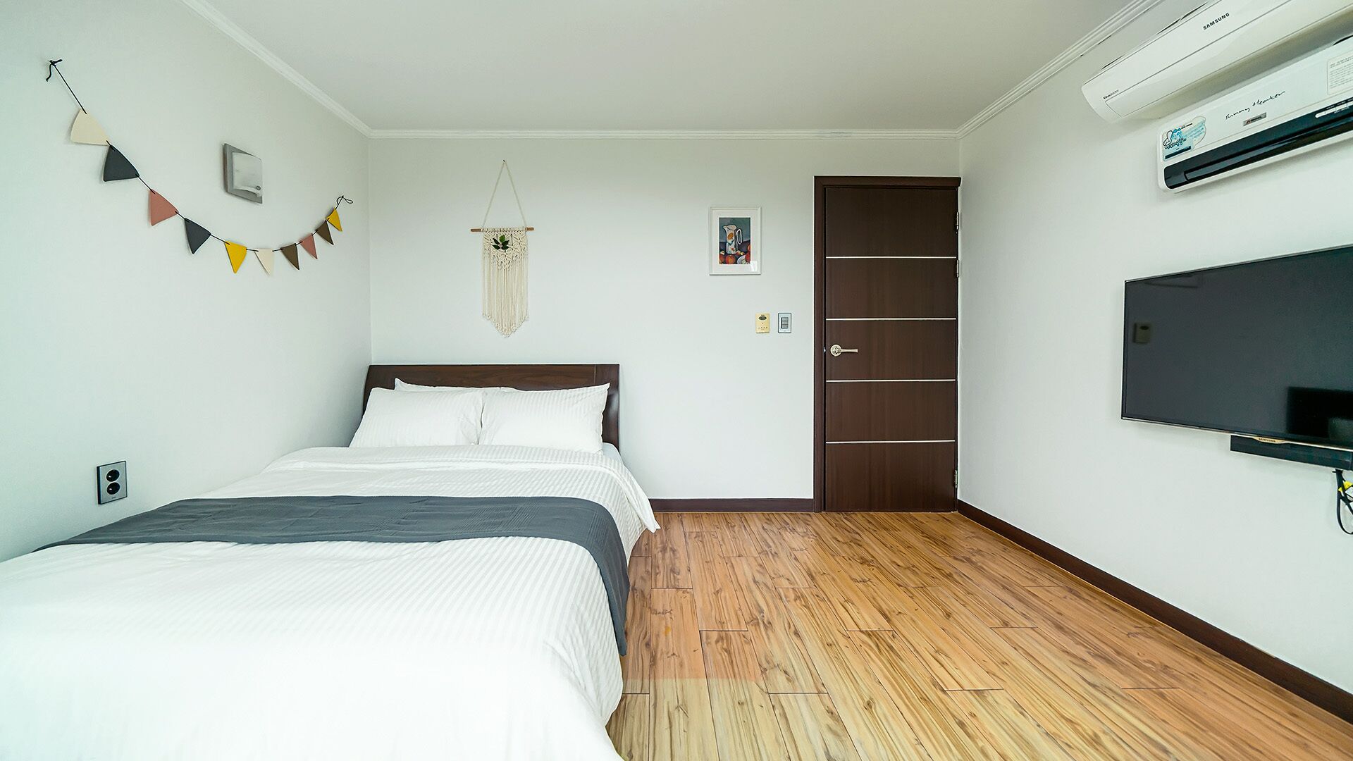 A bedroom with a bed, wooden flooring, a wall-mounted TV, and a door.