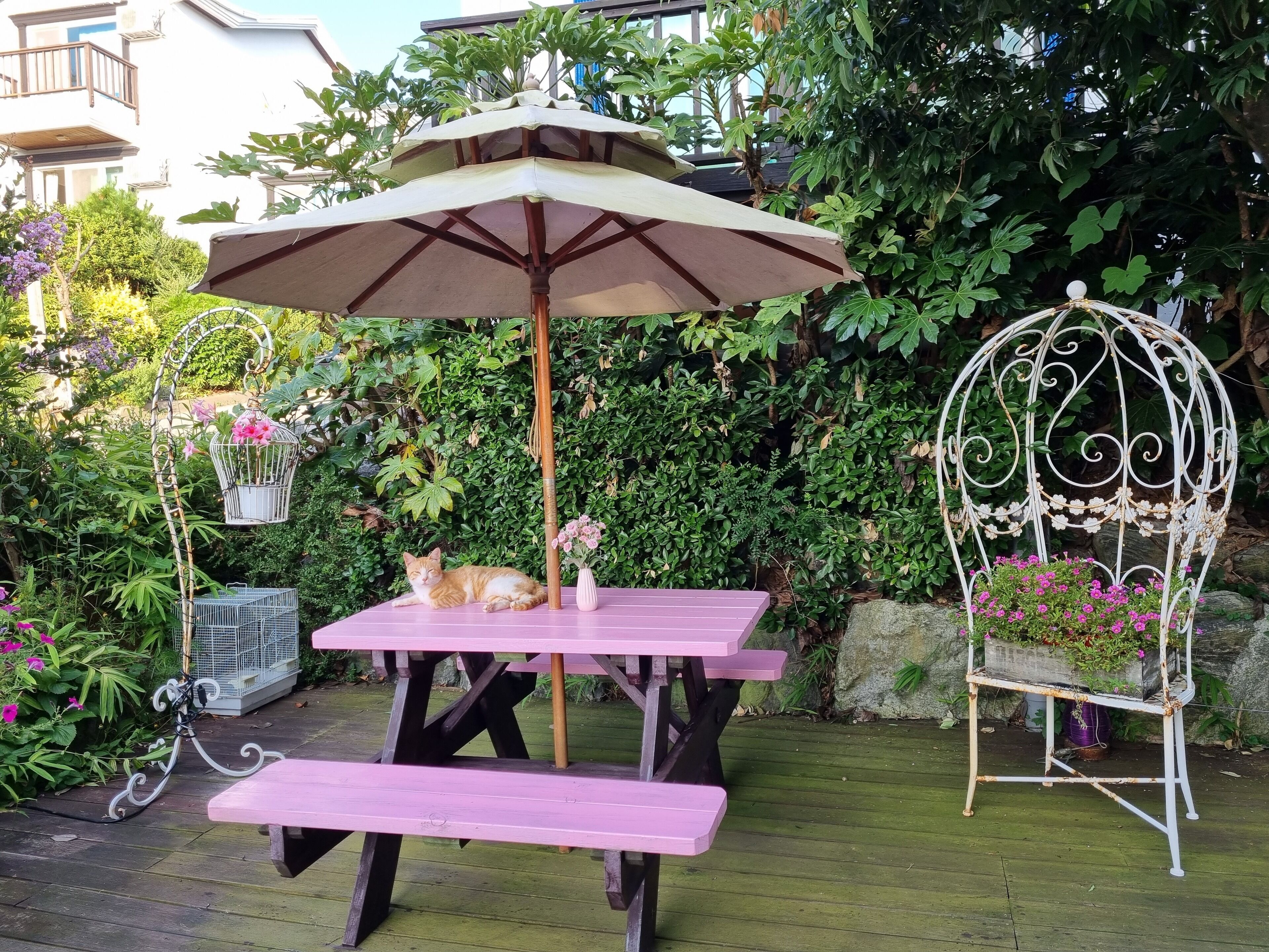 A garden patio with a wooden table and bench, surrounded by lush greenery and a white birdcage with flowers.