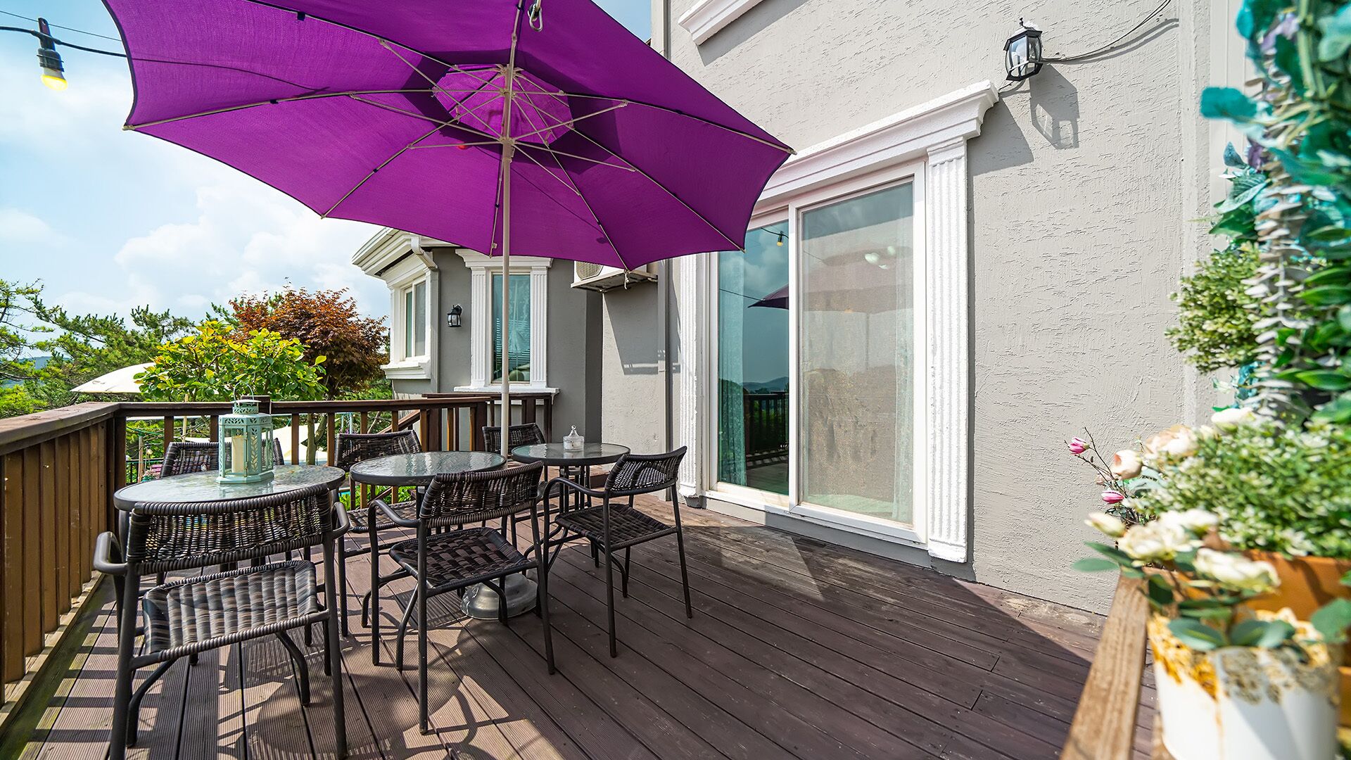 Une terrasse en bois avec des meubles d’extérieur, un parasol violet et une porte vitrée donnant sur un patio.