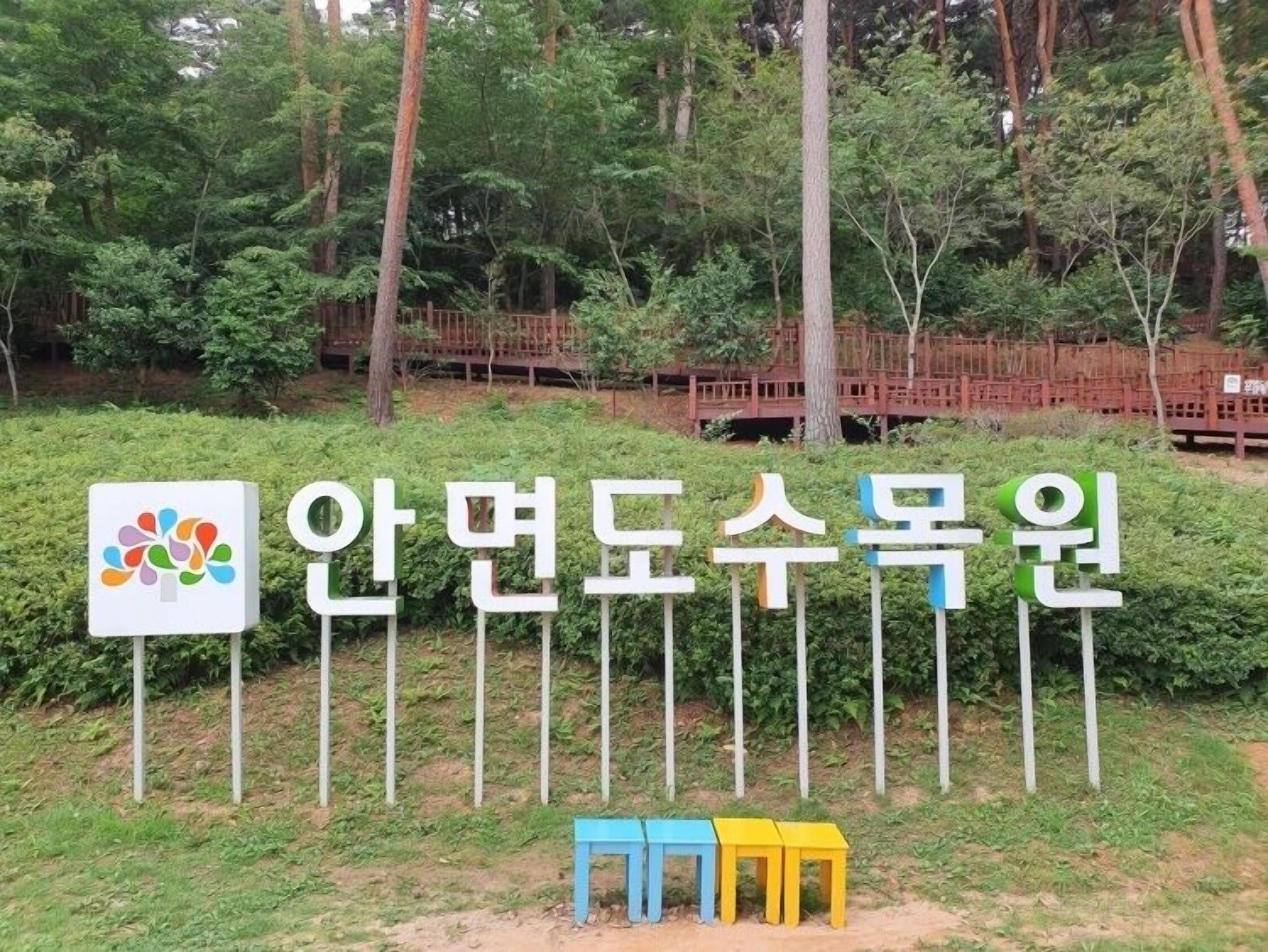 A sign with Korean text and a colorful logo, surrounded by greenery and a wooden fence.