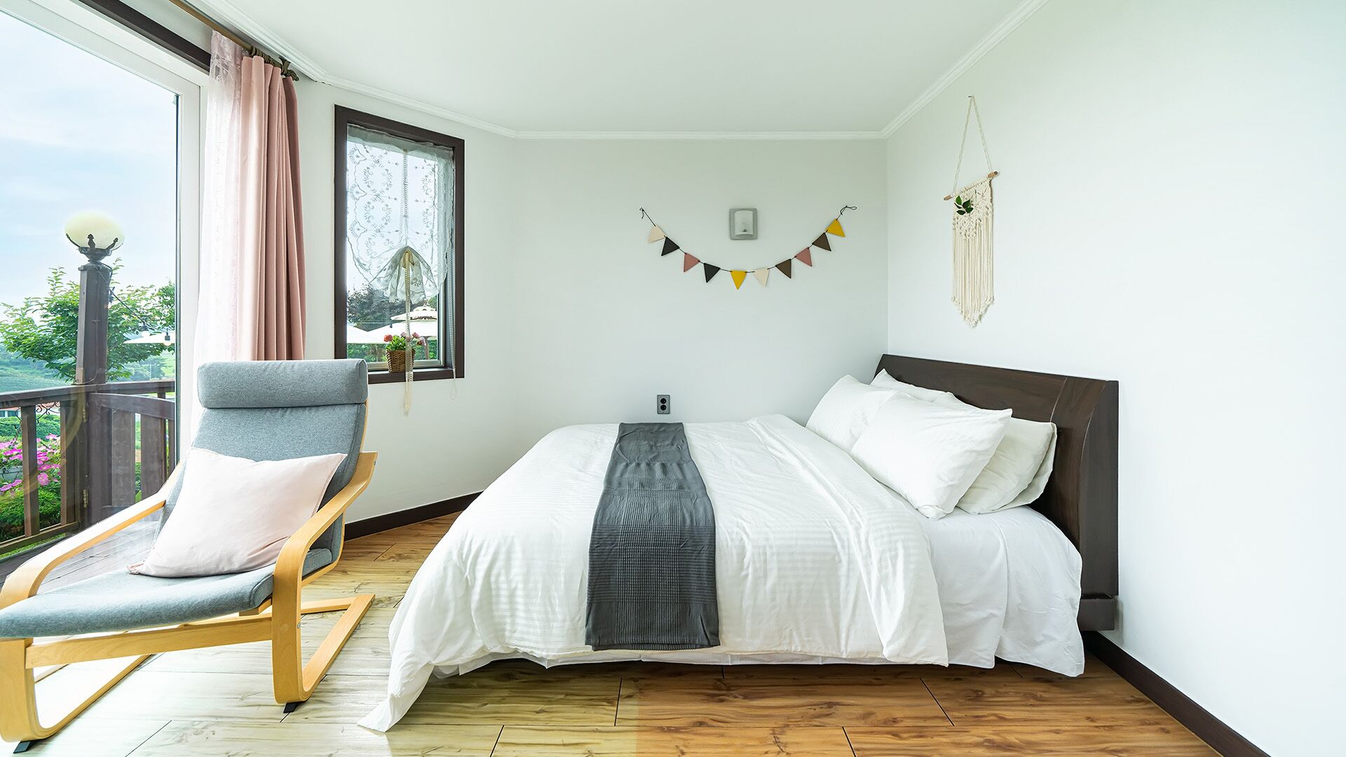 A bedroom with a bed, a chair, a macrame wall hanging, and a view of greenery outside.