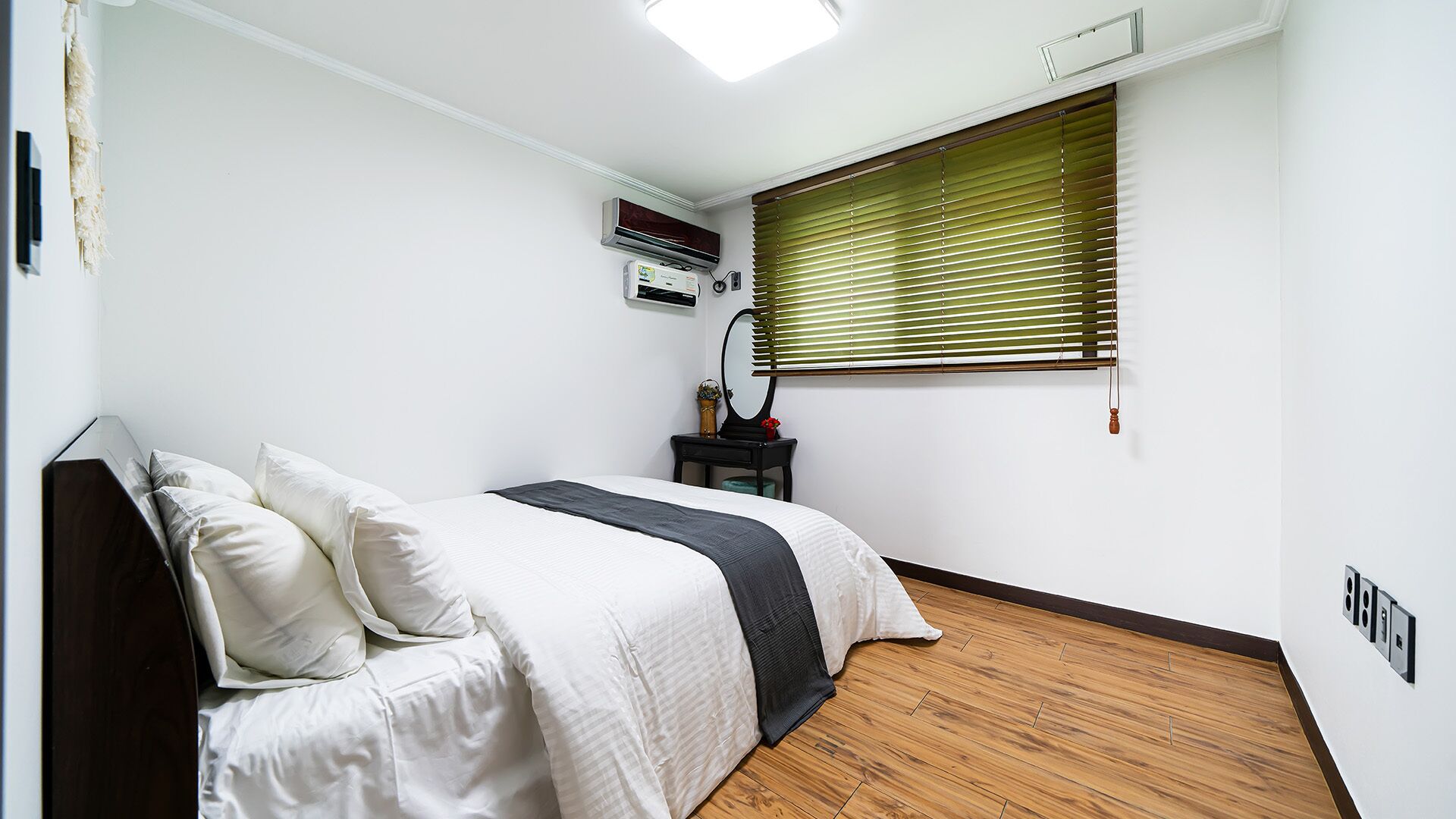 A bedroom with a bed, a nightstand, a mirror, an air conditioner, and a window with blinds.