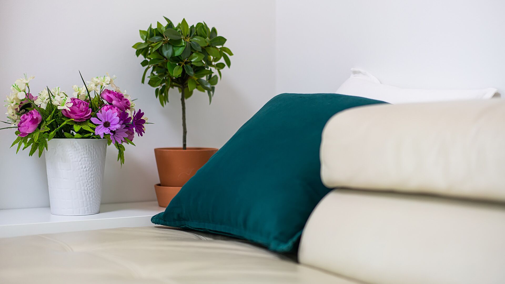 Ett rum med en vit soffa, en turkosfärgad kudde, en vit, strukturerad vas med blommor och en krukväxt.