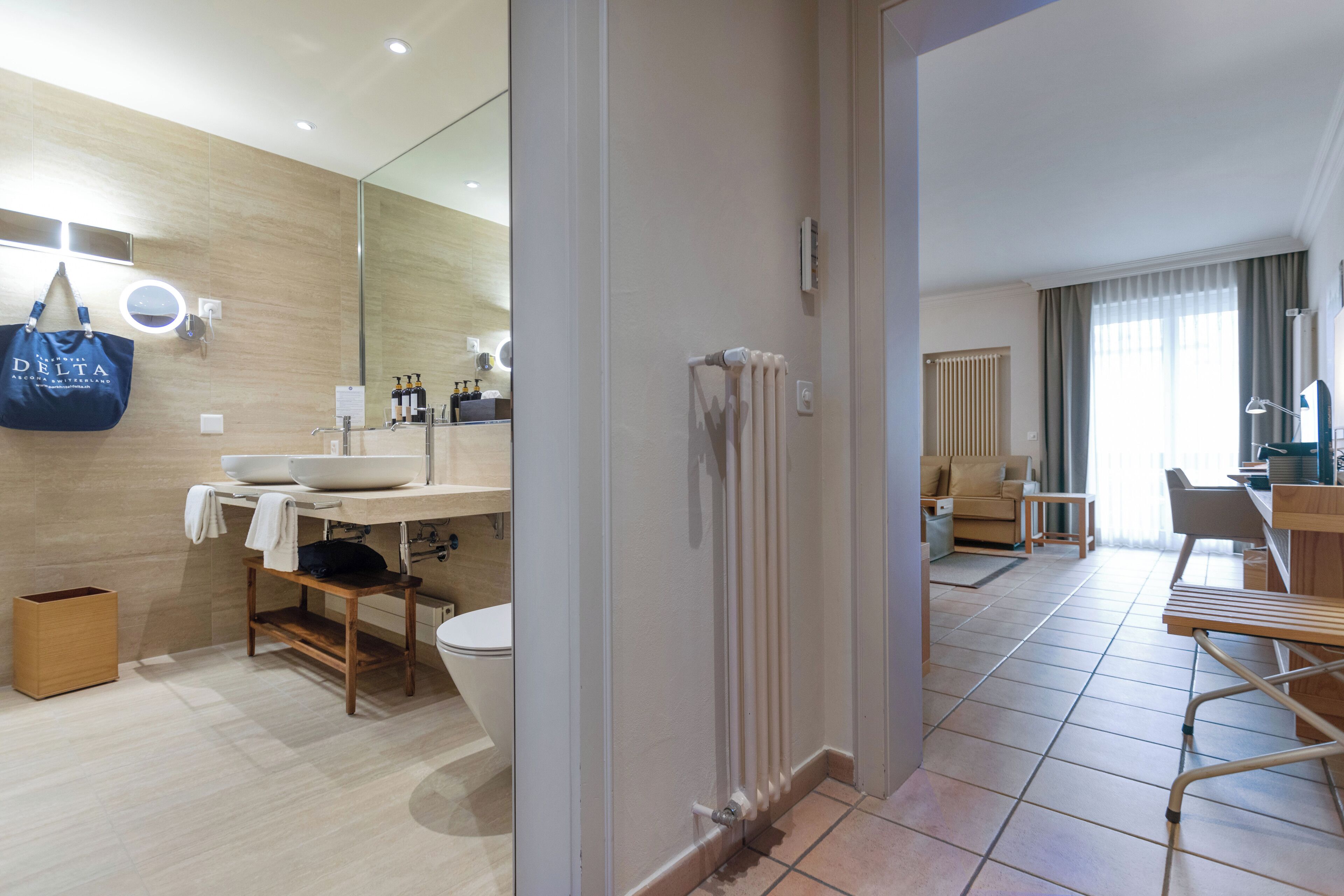 A modern hotel bathroom with a large mirror, sink, and toilet. There is a wooden bench and a table in the adjacent living area.