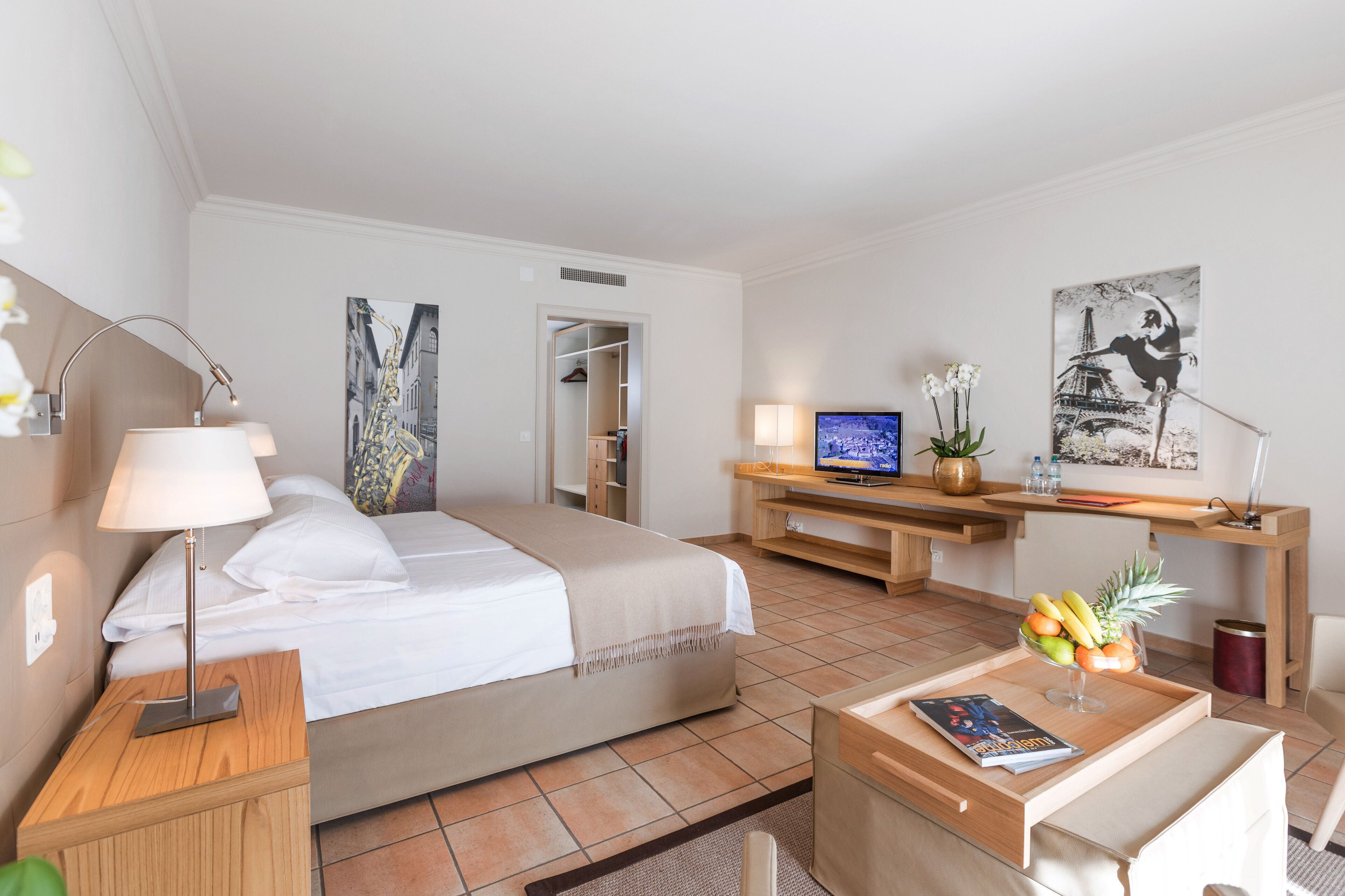 A hotel room with a bed, bedside table, lamp, TV, desk, chair, and artwork.