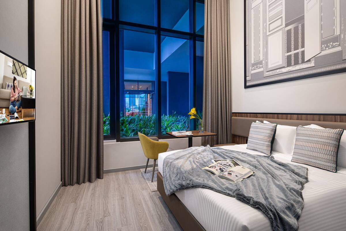 A modern hotel room with a large bed, a desk, a chair, and a large window with curtains.