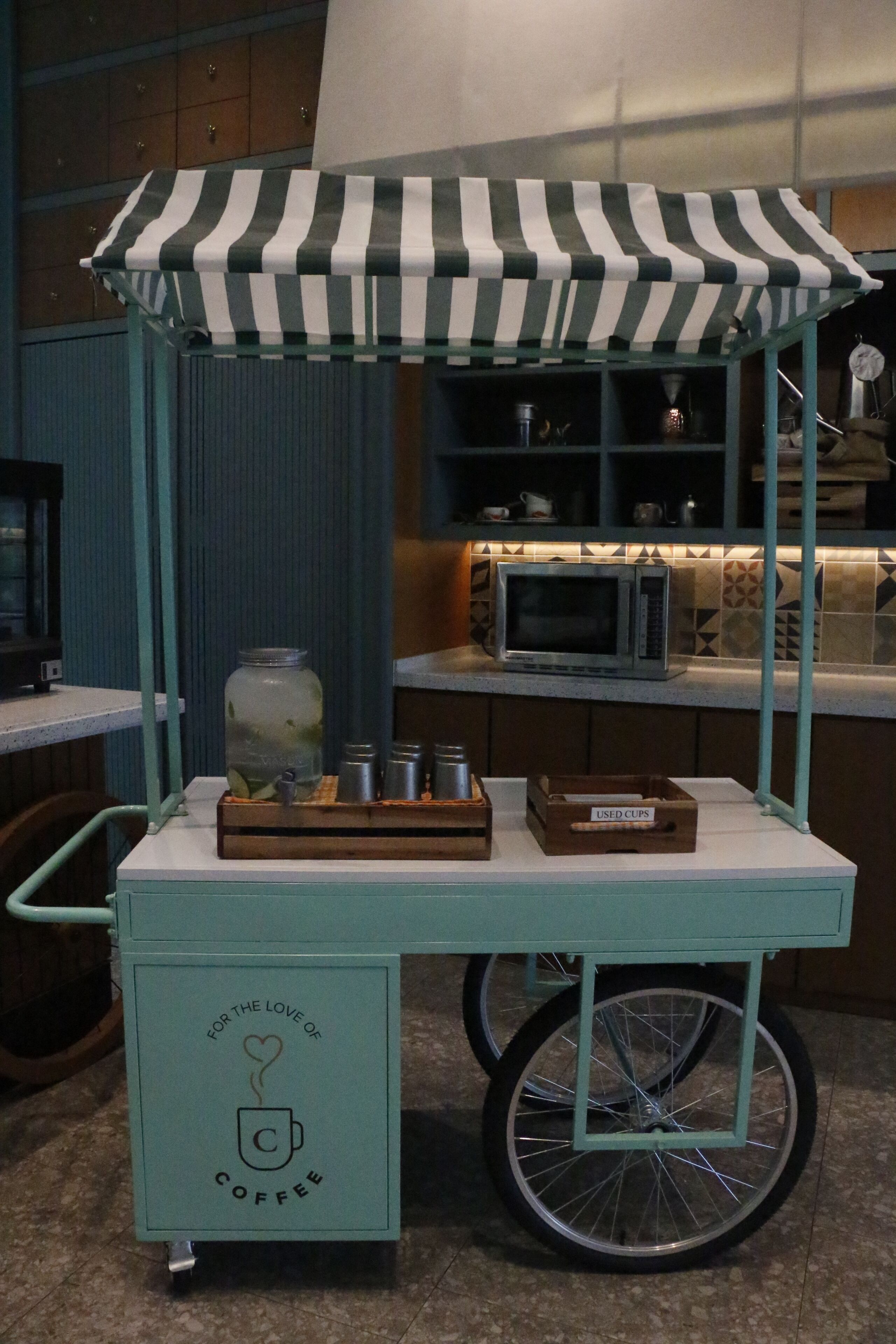 A coffee cart with a striped canopy, a jar of coffee, and a box labeled "USED CUPS" in a kitchen setting.
