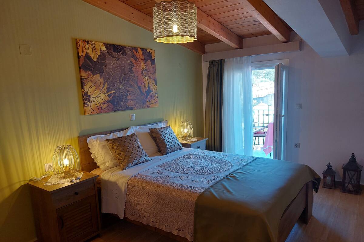 Een slaapkamer met een groot bed, twee nachtkastjes met lampen, een houten hoofdbord, een bloemstil leven op de muur, en een uitzicht op het buitengebeuren door een raam.