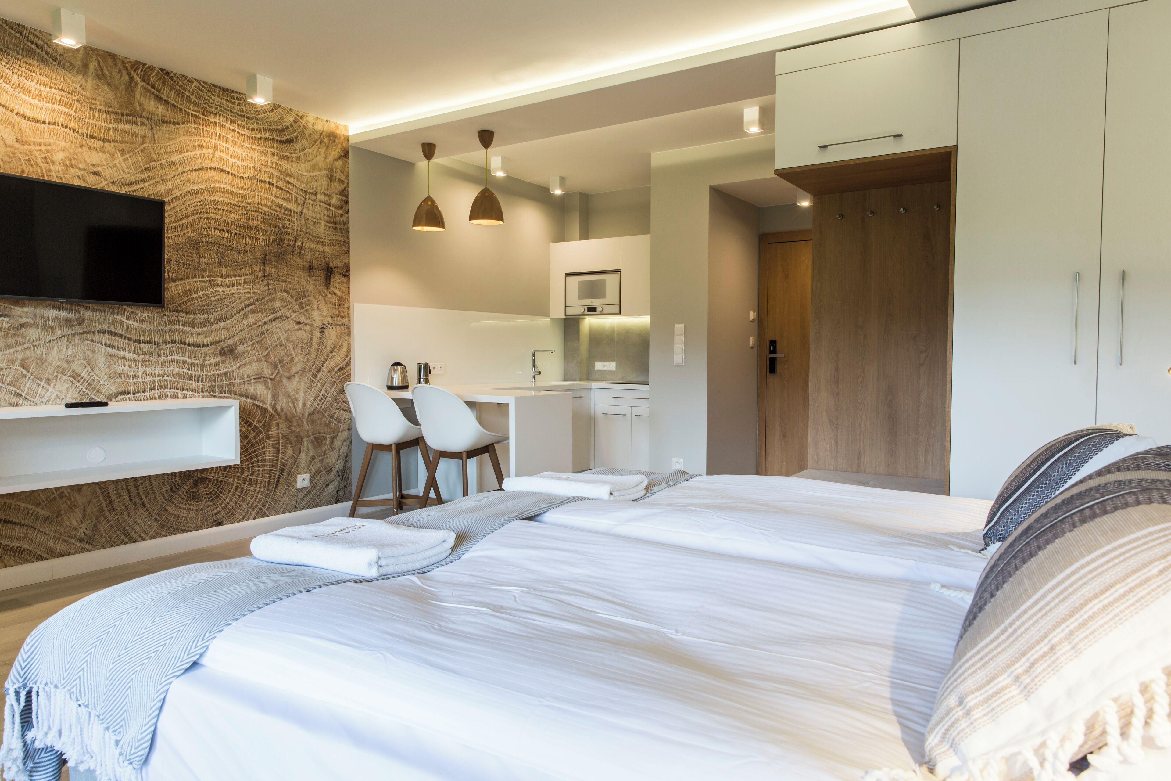 A modern hotel room with a large bed, a TV mounted on a textured wall, a kitchenette, and a wooden wardrobe.