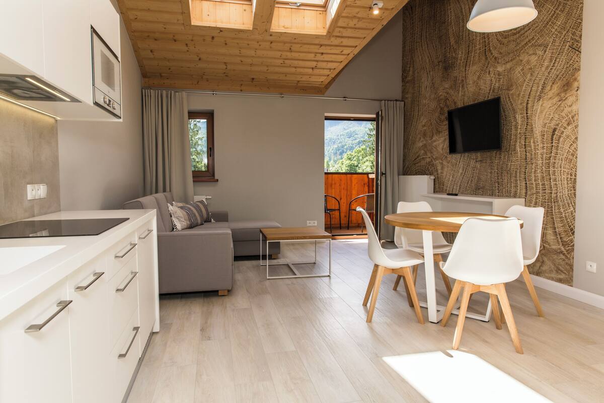 Una cucina moderna con mobili bianchi, un'area pranzo con tavolo in legno e sedie bianche, un divano grigio e una TV montata su una parete strutturata.