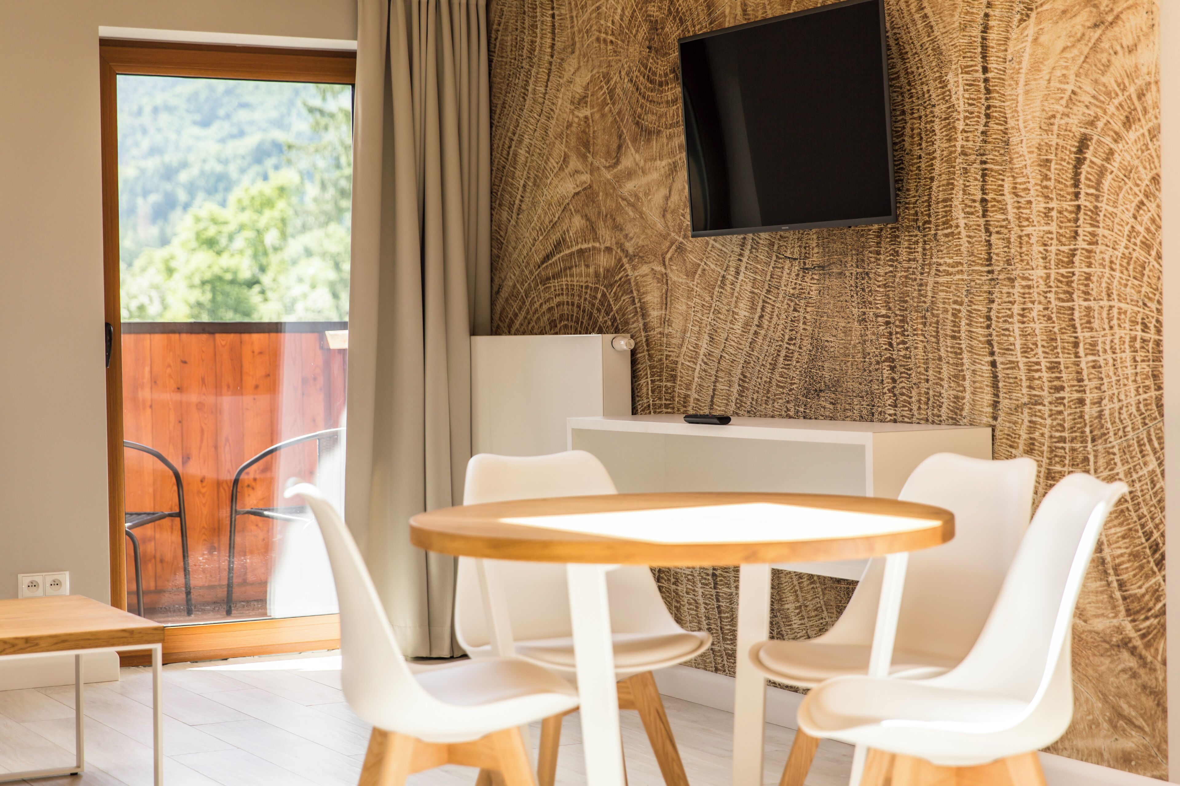 Quarto moderno com uma mesa de madeira e cadeiras brancas, uma TV fixada numa parede texturizada e vista para o verde através de uma janela.