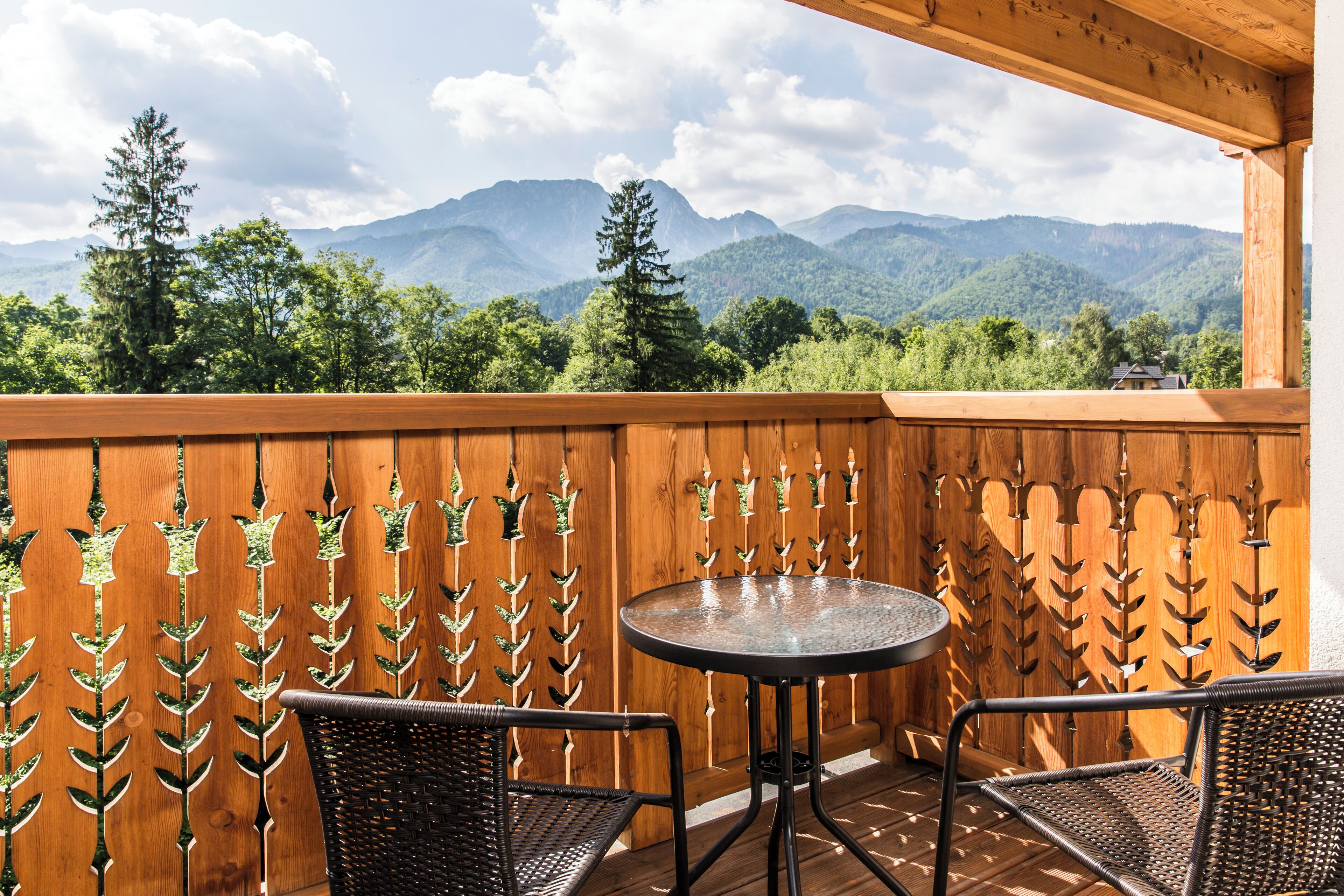 Un balcón de madera con dos sillones de mimbre y una mesa redonda, con vistas a un paisaje montañoso.