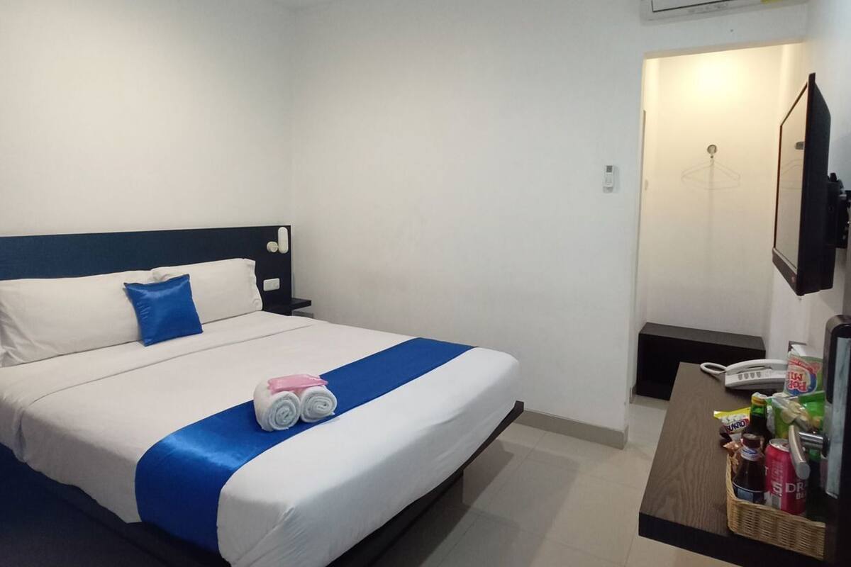 A hotel room with a bed, a bedside table, a desk with a phone, and an air conditioning unit.