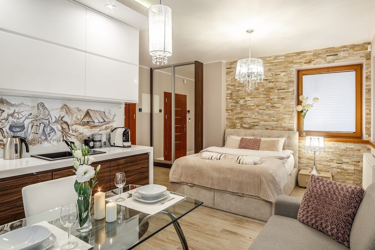 A modern hotel room with a kitchen area, a dining table with a vase of flowers, a bed with a view of the outside, and a chandelier.