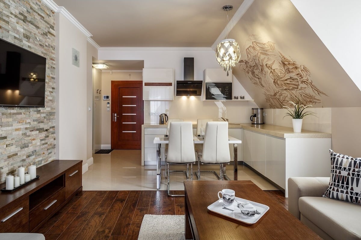 A modern living room with a stone wall, wooden flooring, a flat-screen TV, a dining area with white chairs, a kitchen with a stove and microwave, and a staircase with a decorative wall piece.