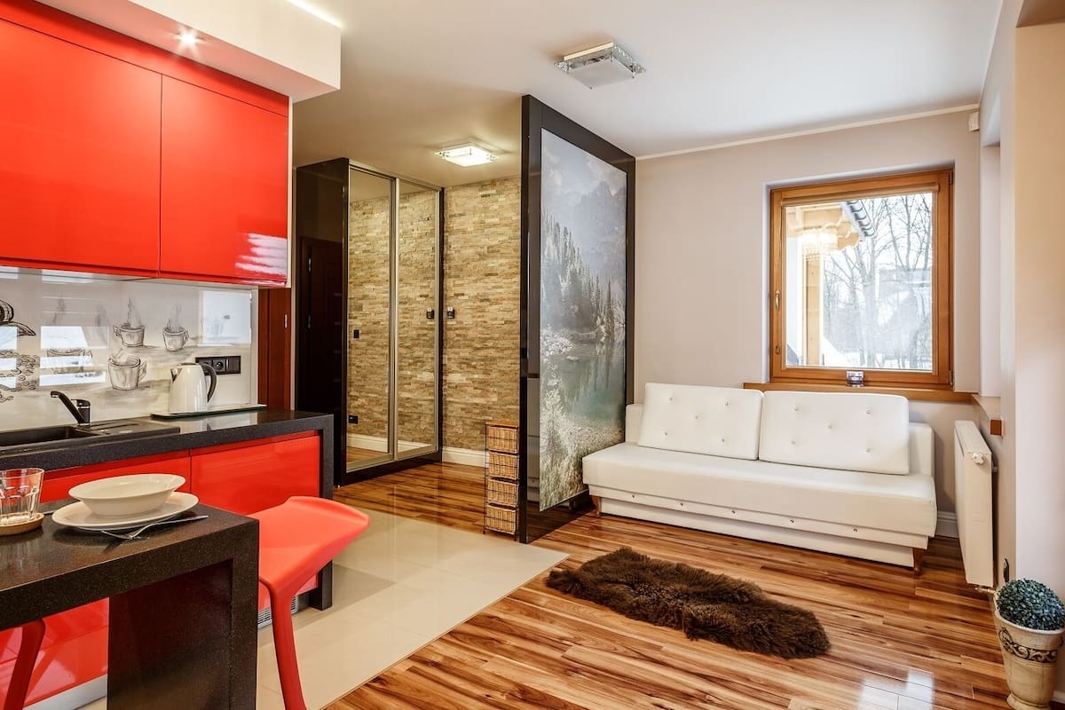A modern kitchen with red cabinets, a dining area with a white table and red chair, a white sofa, and a large window.