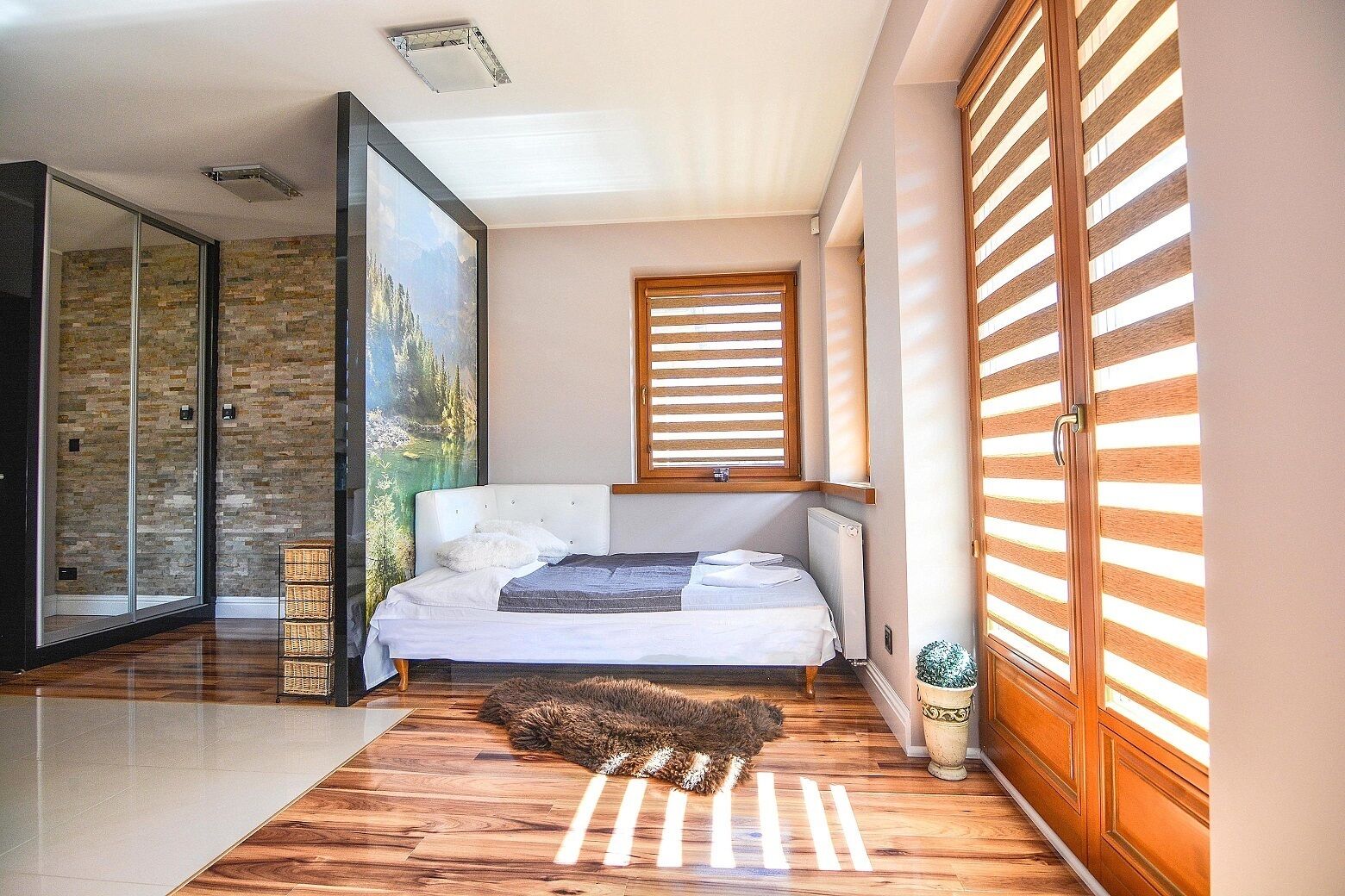 A bedroom with a bed, a large window with blinds, a stone wall, and a sliding glass door.