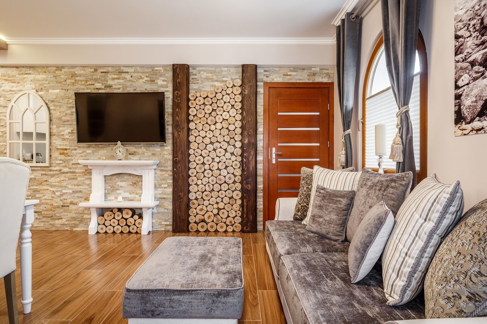 A living room with a stone wall, a TV mounted above a fireplace, a wooden door, and a sofa with patterned cushions.