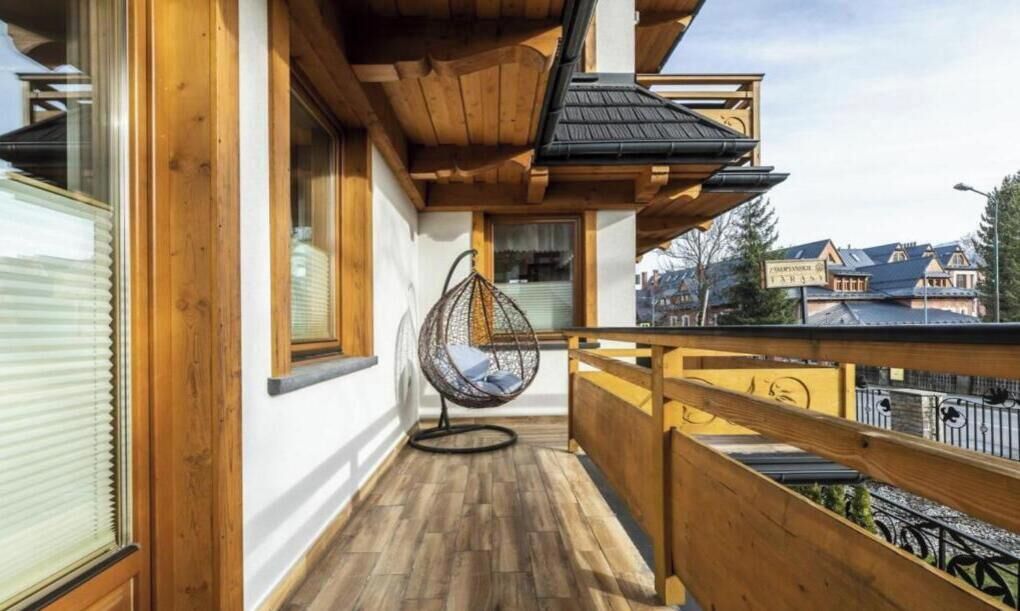 Een houten balkon met een hangstoel, houten leuningen en een uitzicht op gebouwen.