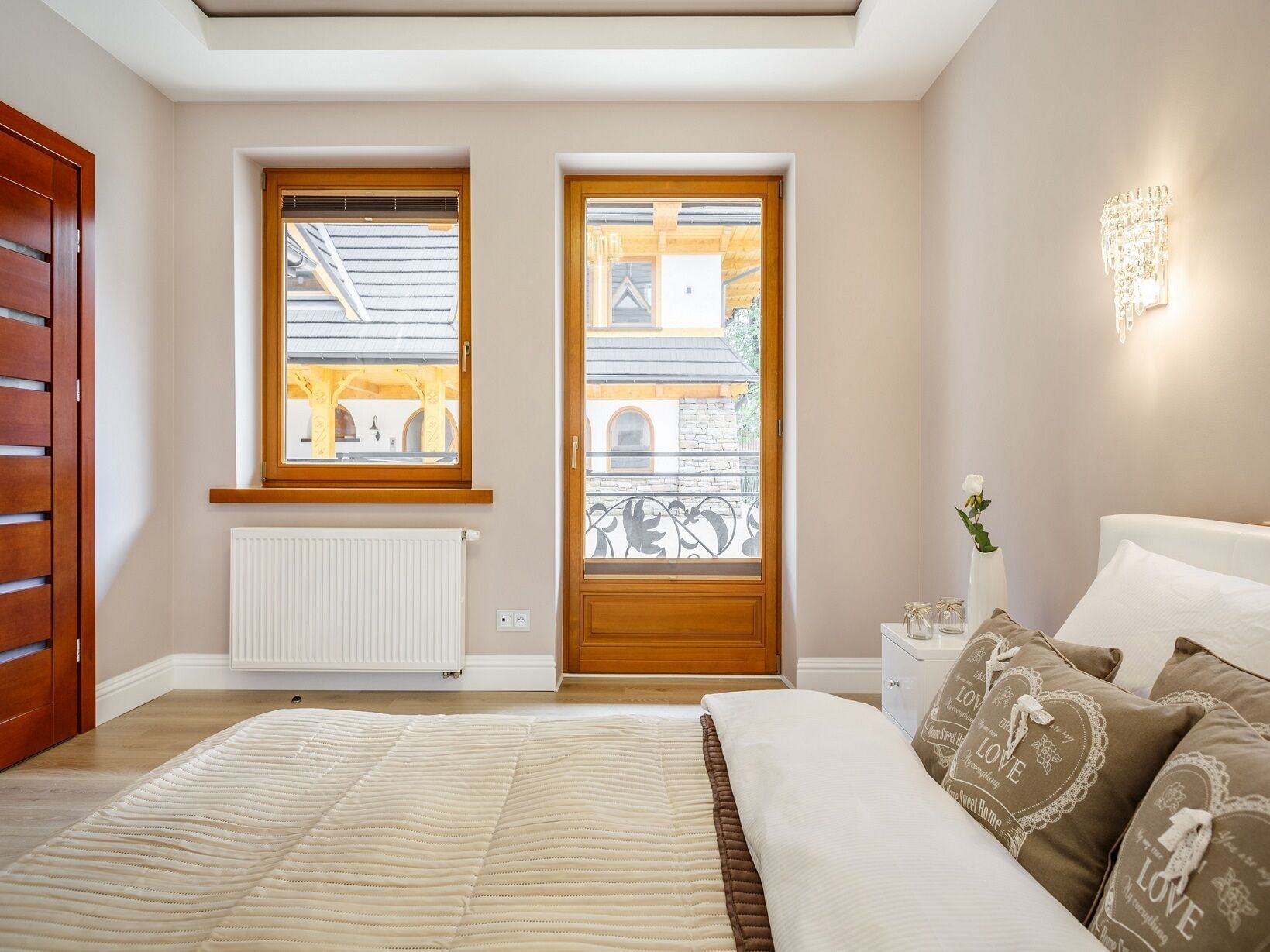 A bedroom with a large bed, two windows, a radiator, a bedside table with a vase, and a wall-mounted light fixture.