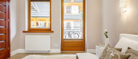 A bedroom with a large bed, two windows, a radiator, a bedside table with a vase, and a wall-mounted light fixture.