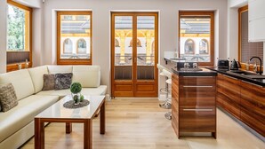 A modern kitchen with wooden cabinets, a black countertop, and a sink. There is a dining area with a white table and a sofa. Large windows provide views of a building with arches.