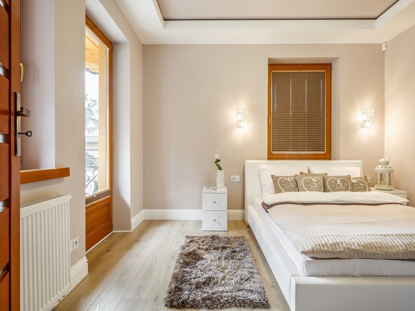 A bedroom with a bed, bedside tables, a nightstand, a window with blinds, and a radiator.