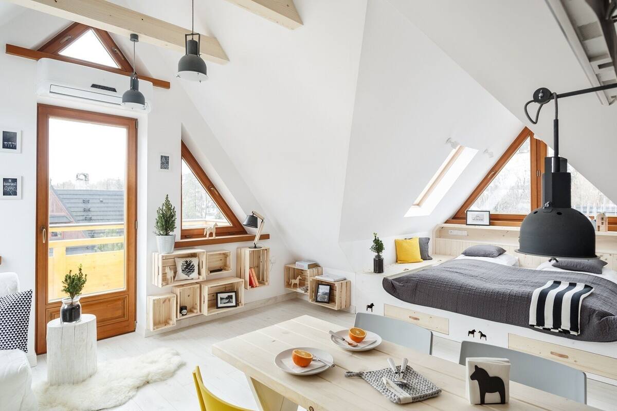 A high-ceilinged attic bedroom with a sloped roof, wooden beams, a skylight, a bed with a gray blanket, a desk with breakfast items, and a view of the outside.