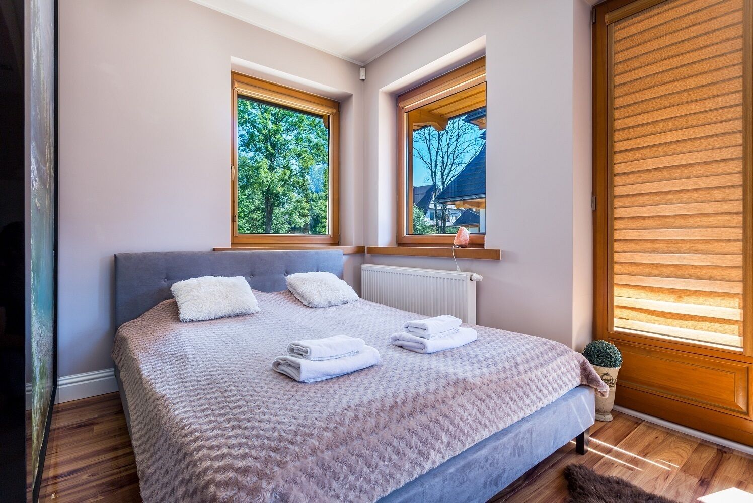 A bedroom with a bed, two windows, a radiator, and a small plant.