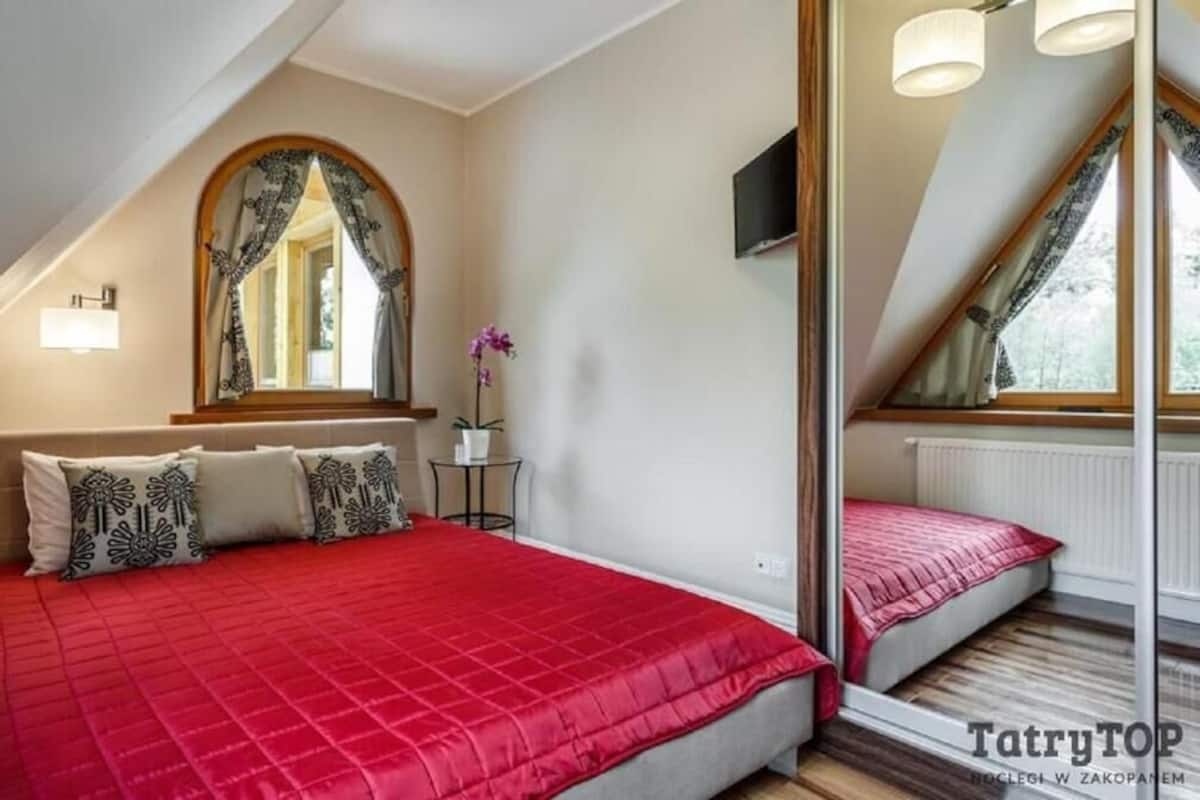 A bedroom with a red bedspread, a mirror, a small table with a potted plant, and a window with curtains.