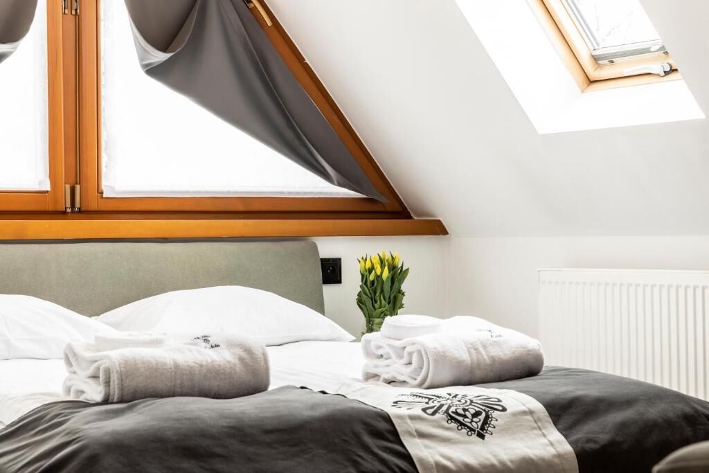 Une chambre d’hôtel avec un lit, une fenêtre avec des rideaux, un radiateur et un vase rempli de fleurs.