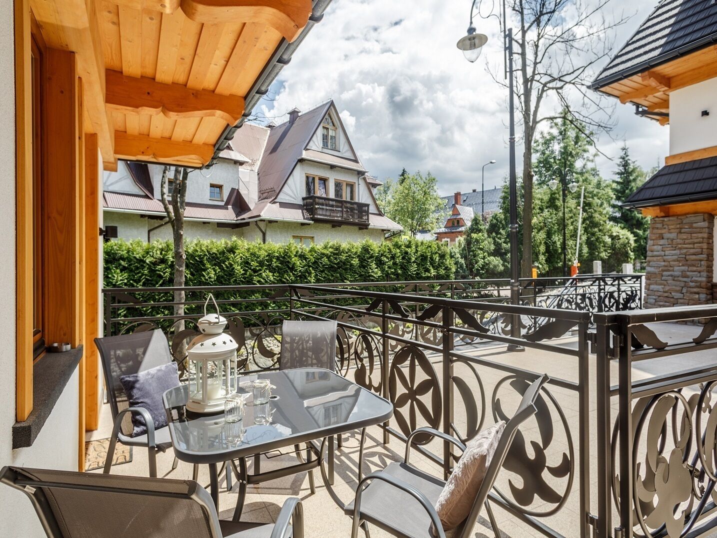 A balcony with a table and chairs, a lantern, and a view of houses in the background.