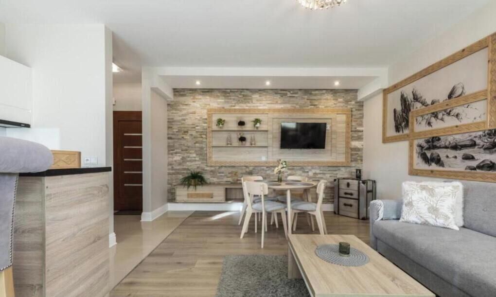 A modern living room with a stone accent wall, a flat-screen TV, a dining area with a table and chairs, and a gray sofa.