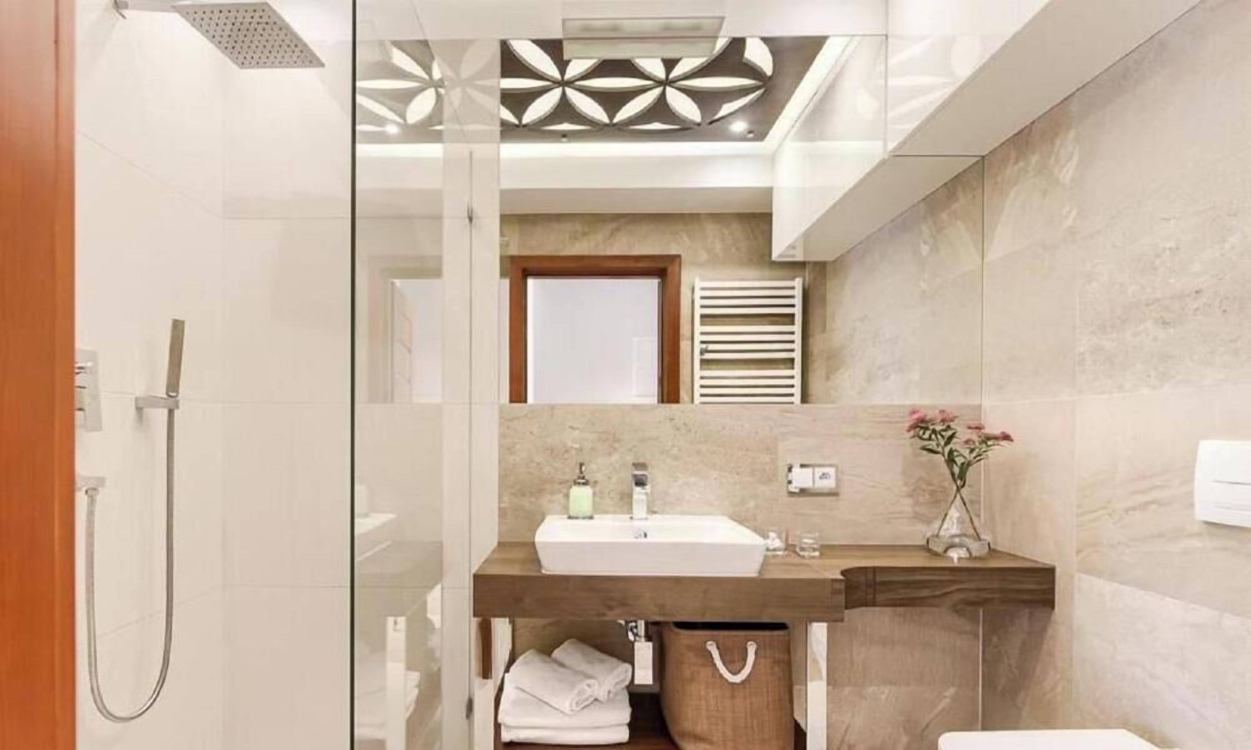 Un baño moderno con cabina de ducha de vidrio, tocador de madera, pared con azulejos de mármol y un panel decorativo en el techo.