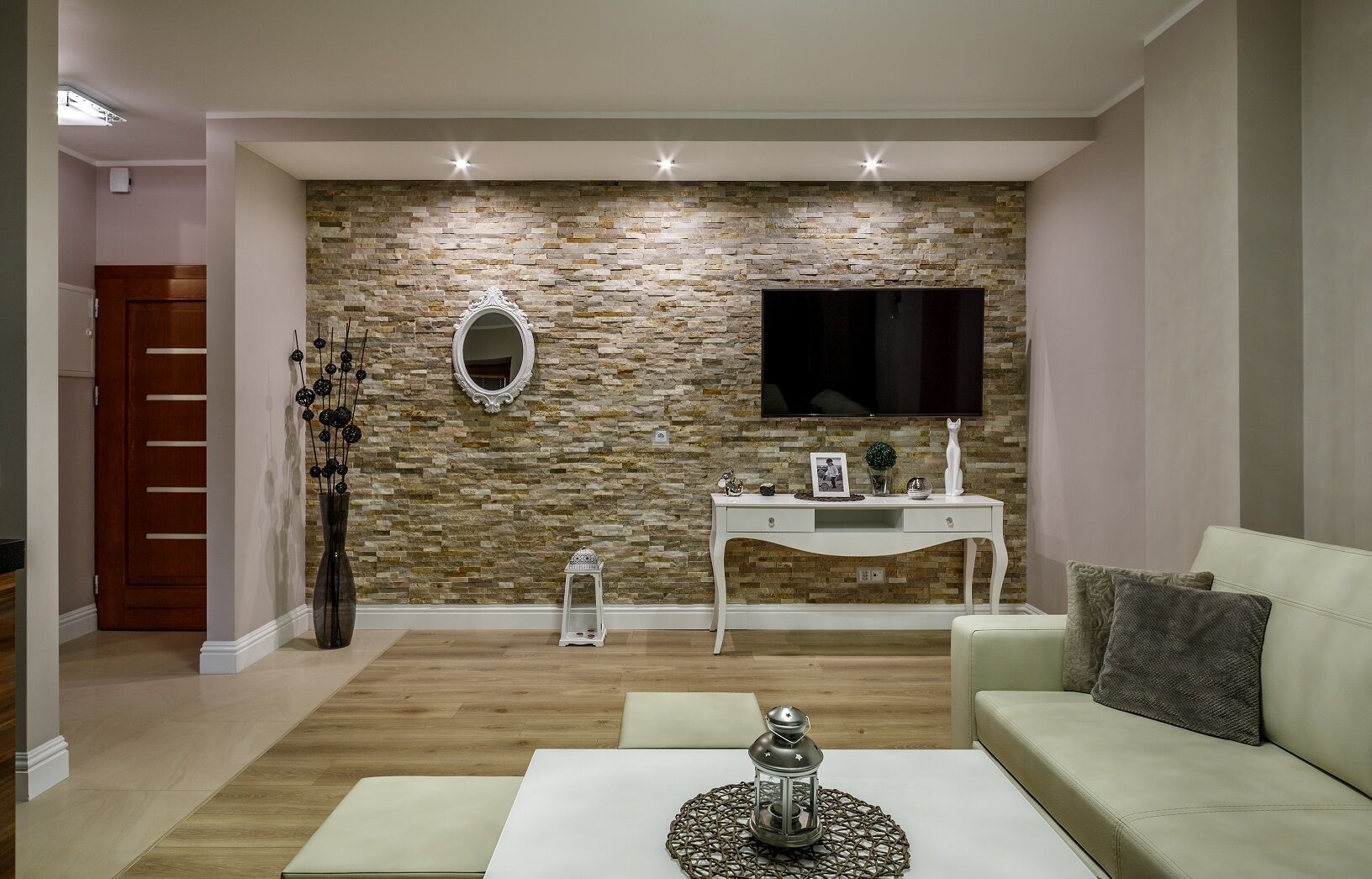A modern living room with a stone wall, a white console table, a flat-screen TV, a white sofa, and a coffee table with a decorative item.