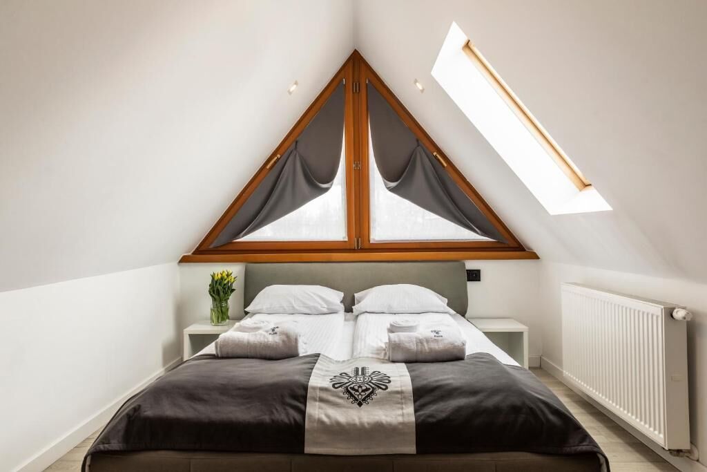 A bedroom with a sloped ceiling, a bed with a dark headboard, a bedside table with a vase of flowers, and a window with curtains.